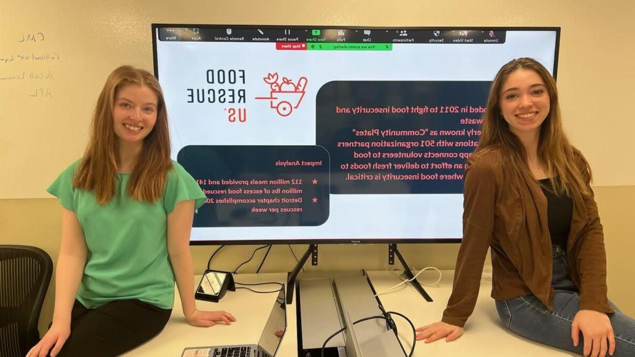 十大菠菜台子学生, 莎拉·奈奎斯特和瑞秋·惠伦, 坐在一个大显示器前，上面显示着美国食品救援组织的信息.